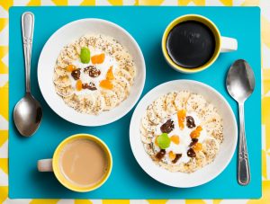 Top view of two breakfast bowls with bananas, yogurt, and coffee cups on a vibrant background.